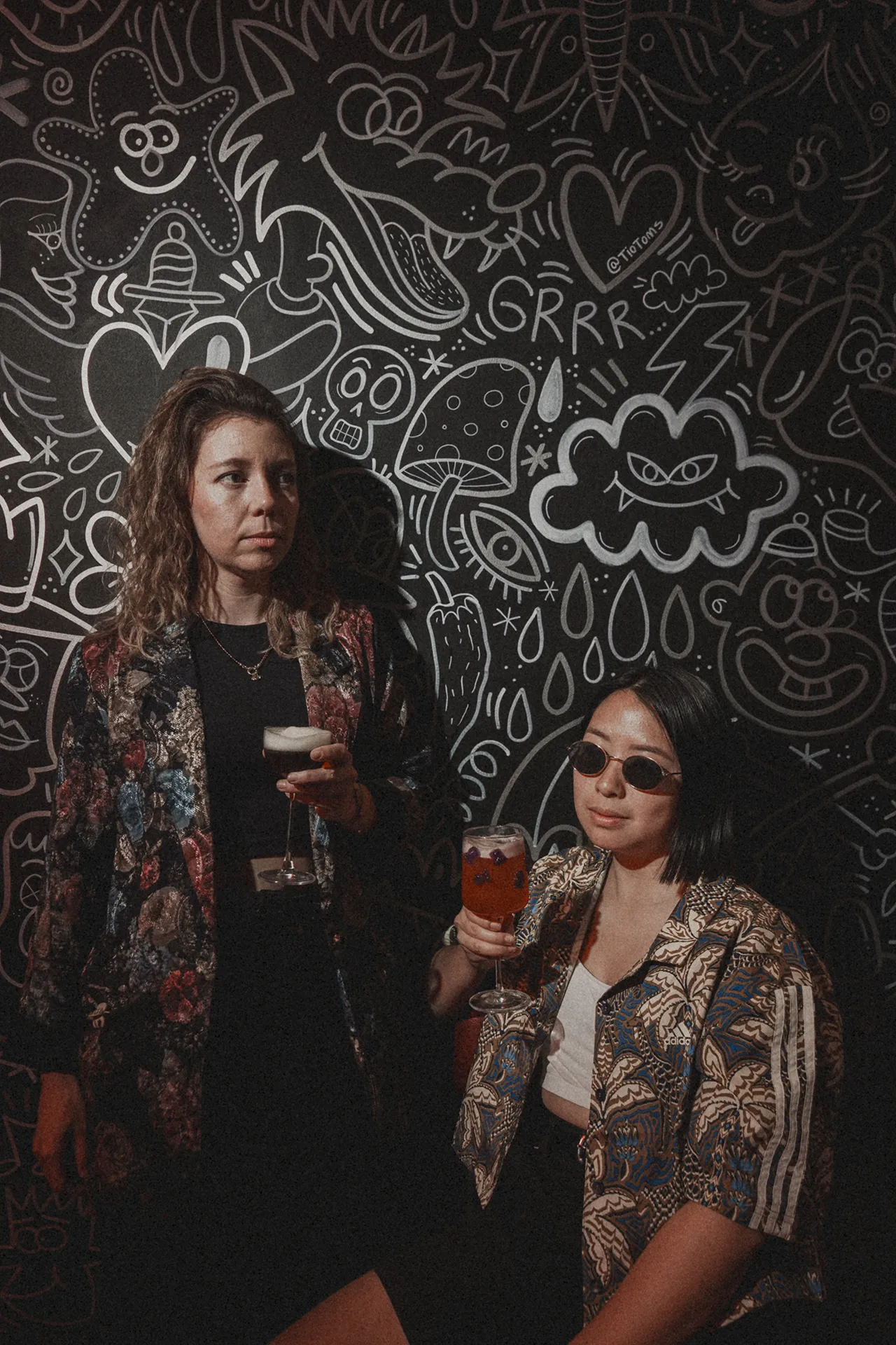 Dos amigas con cócteles frente al mural ilustrado de Prima